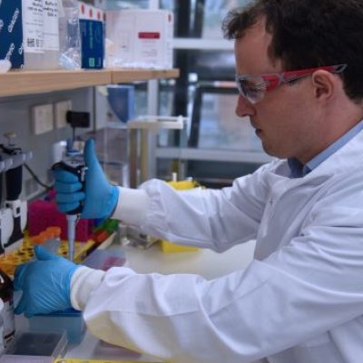 A man in a white lab coat, goggles and gloves in profile, working at a laboratory bench.